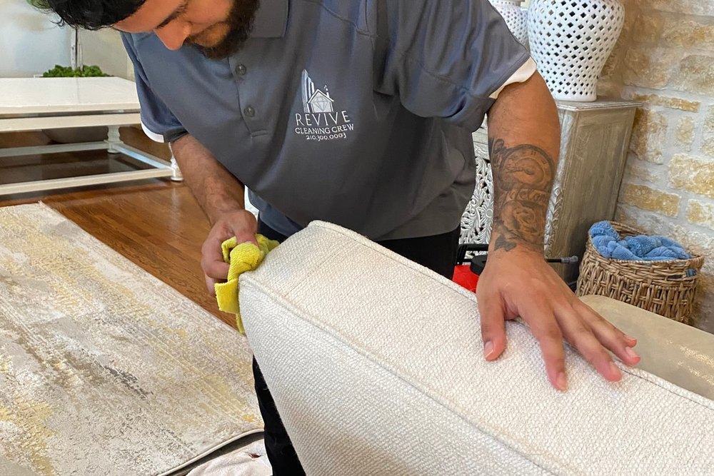 A Revive Cleaning Crew technician cleaning the arm of a cream upholstered sofa by hand in a San Antonio home