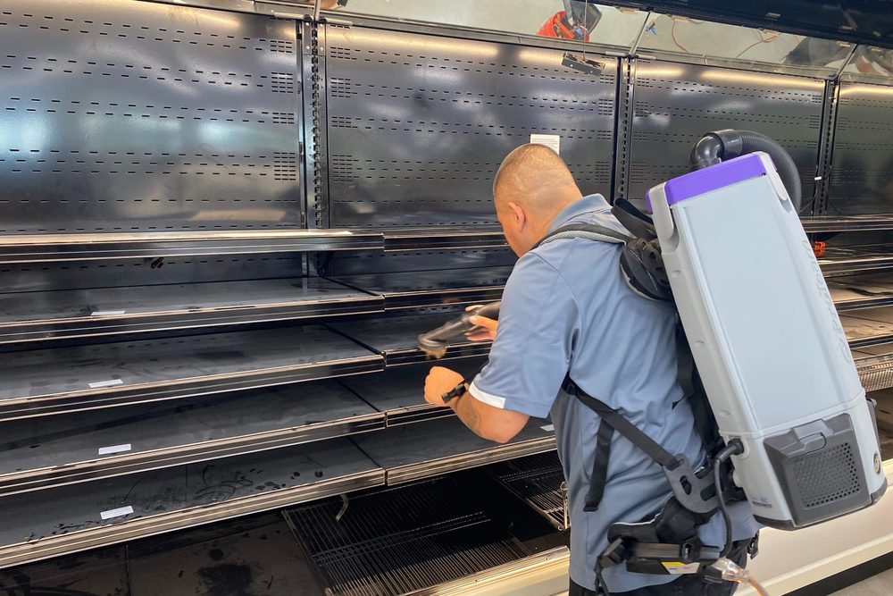 A Revive technician wearing a backpack vacuum cleaning the shelves of an empty refrigerated display case